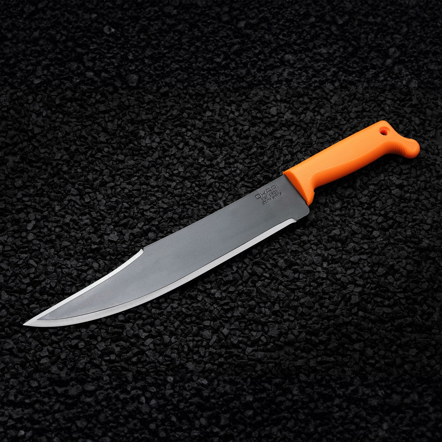 Okapi Bowie-style machete with Okapi Orange grip resting on black stone ground