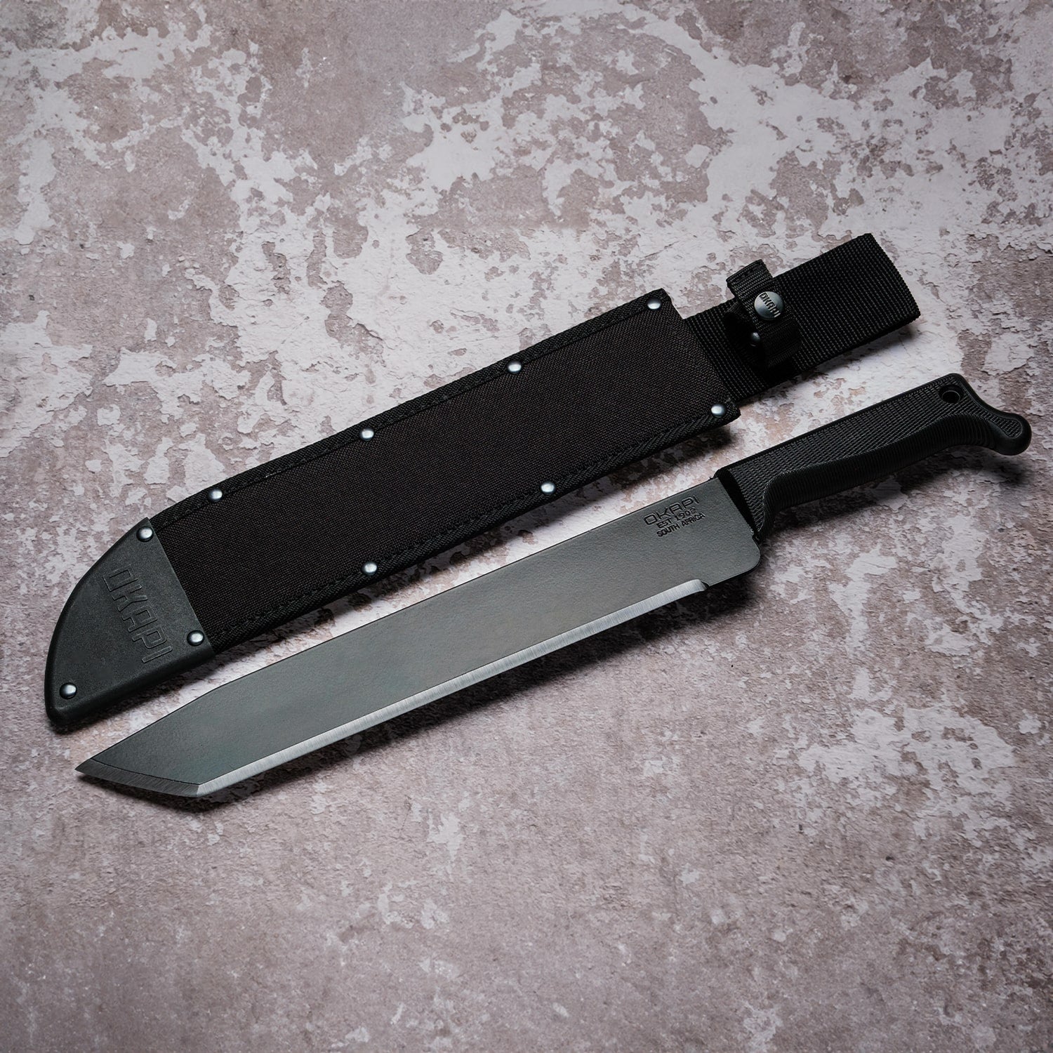 Okapi O-Tanto machete with canvas sheath resting on rugged grey stone floor