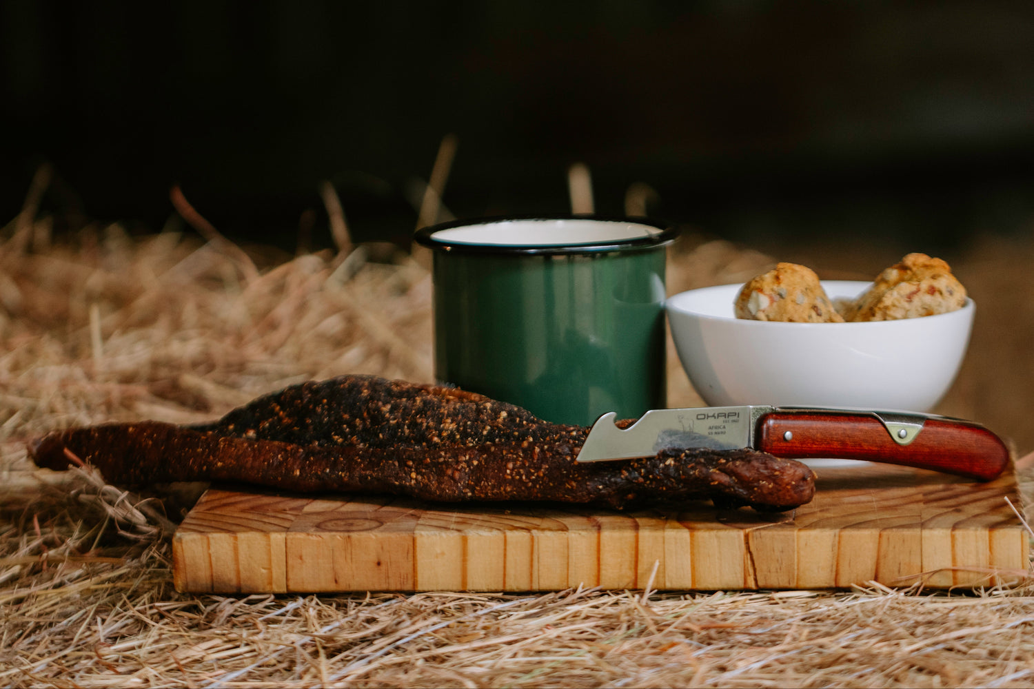 Meat on a wooden cutting board with a knife, green mug, and white bowl on hay.
